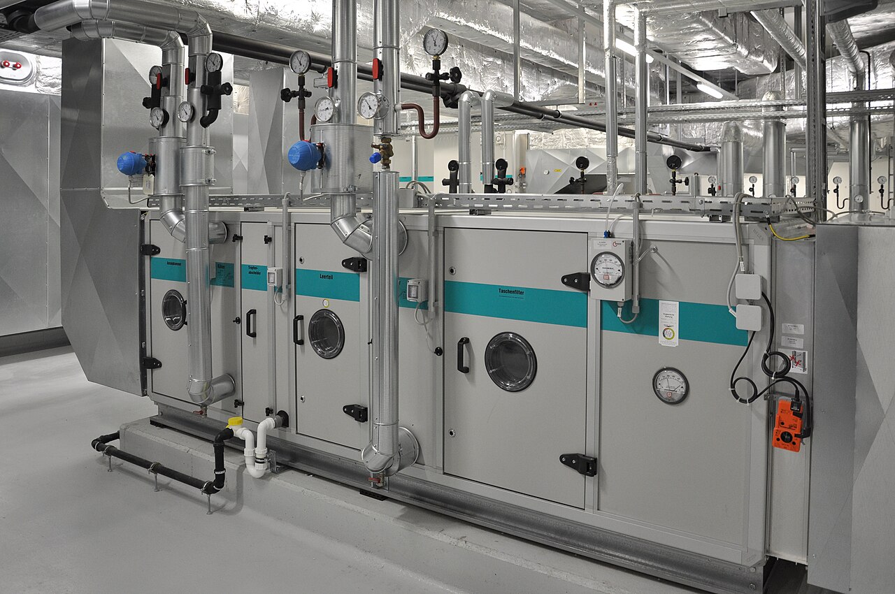 Centrale de traitement d'air dans un local technique : longs caissons gris à hublots, raccordés à des tuyaux munis de vannes et de thermomètres